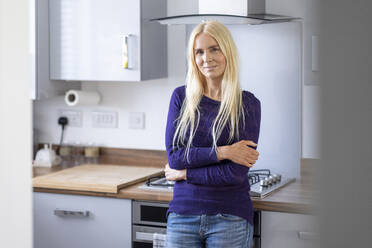 Blond woman with arms crossed leaning on kitchen counter at home - WPEF05310