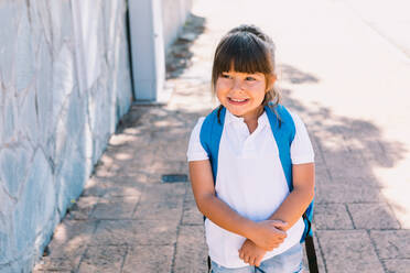 Fröhliches Schulkind mit braunen Haaren in weißem T-Shirt und mit buntem Rucksack, das auf einem gekachelten Gehweg in der Stadt wegschaut - ADSF30336