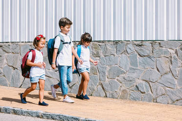 Schoolboy with backpack speaking with female friends while strolling on tiled pavement against stone wall in sunlight - ADSF30338