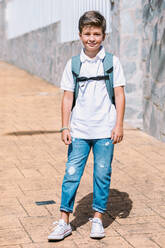 Zufriedenes Schulkind in zerrissenen Jeans und Gummistiefeln schaut in die Kamera auf einem gekachelten Bürgersteig in einer sonnigen Stadt - ADSF30340