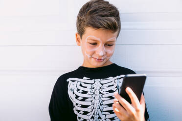 Cheerful boy in black skeleton costume with painted face using on mobile phone while sitting near white wall on street - ADSF30349