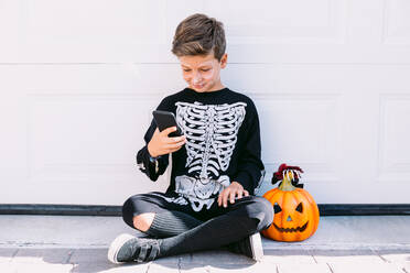 Full body of cheerful boy in black skeleton costume with painted face and carved Halloween pumpkin using on mobile phone while sitting near white wall on street - ADSF30350