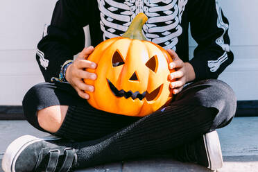 Crop unrecognizable child in black skeleton costume holding creepy carved Halloween Jack O Lantern pumpkin while sitting with legs crossed on street - ADSF30352