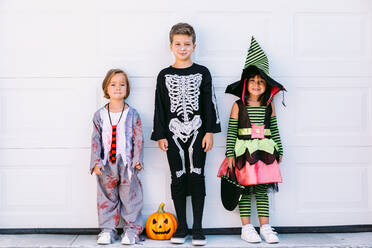 Full body of group of little kids dressed in various Halloween costumes with carved Jack O Lantern standing near white wall on street - ADSF30354