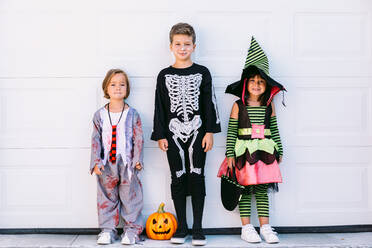 Ganzkörpergruppe von kleinen Kindern in verschiedenen Halloween-Kostümen mit geschnitzter Jack O Lantern, die in der Nähe einer weißen Wand auf der Straße stehen - ADSF30354