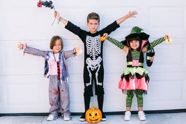 Cheerful little friends in various Halloween costumes with pumpkin and accessories raising arms and looking at camera while standing together near white wall - ADSF30355