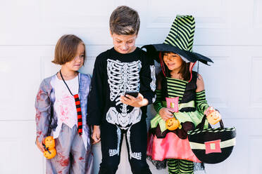 Full body of group of little kids dressed in various Halloween costumes with carved Jack O Lantern browsing mobile phone together while standing near white wall on street - ADSF30356