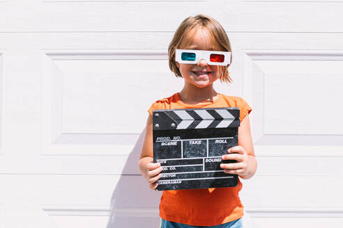 Cute happy girl in casual colorful clothes holding clapperboard while on three dimensional glasses standing on white wall background - ADSF30357