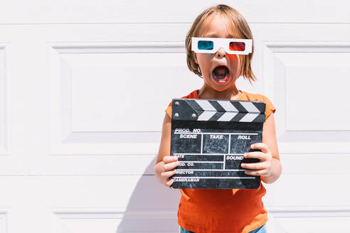 Cute astonished girl in casual colorful clothes holding clapperboard while on three dimensional glasses standing on white wall background - ADSF30358
