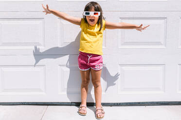 Fröhliches süßes Mädchen in lässiger bunter Kleidung und dreidimensionaler Brille auf weißem Wandhintergrund stehend - ADSF30359