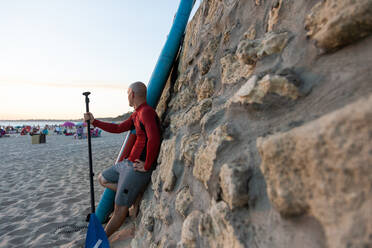 Seitenansicht eines männlichen Surfers in Neoprenanzug und Hut, der mit Paddel und SUP-Board am Strand steht und sich an eine Steinmauer lehnt, um zu surfen - ADSF30381