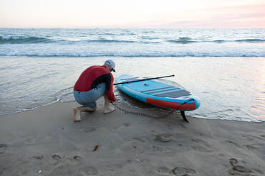 Rückansicht eines männlichen Surfers in einem Neoprenanzug, der die Knöchelleine in das SUP-Board einhängt, während er sich auf das Paddelsurfen am Meeresufer vorbereitet - ADSF30382