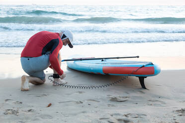 Rückansicht eines männlichen Surfers in einem Neoprenanzug, der die Knöchelleine in das SUP-Board einhängt, während er sich auf das Paddelsurfen am Meeresufer vorbereitet - ADSF30383