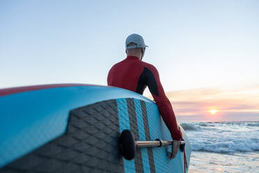 Rückenansicht eines nicht erkennbaren männlichen Surfers in Neoprenanzug und Hut, der ein Paddelbrett trägt und ins Wasser geht, um am Meeresufer zu surfen - ADSF30385