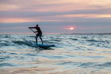 Männlicher Surfer in Neoprenanzug und Hut auf einem Paddelbrett beim Surfen am Meeresufer während des Sonnenuntergangs - ADSF30387