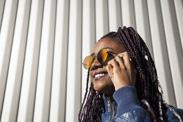 Fröhliche, stilvolle junge Afroamerikanerin mit Afrozöpfen, die eine trendige Jacke und eine Sonnenbrille trägt und mit einem Mobiltelefon spricht, während sie in der Nähe der Wand eines städtischen Gebäudes steht - ADSF30396