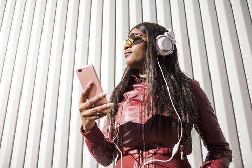 Niedriger Blickwinkel auf eine trendige junge langhaarige Afroamerikanerin in roter Jacke und Sonnenbrille, die über Kopfhörer und Mobiltelefon Musik hört, während sie einen sonnigen Tag in der Stadt genießt - ADSF30397