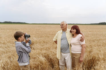 Grandson photographing grandparents through camera in wheat field - EYAF01742