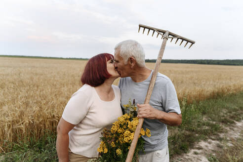 Ehepaar mit Blume und Harke küsst sich auf einem Weizenfeld - EYAF01764