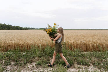 Mädchen hält einen Korb mit Rainfarnblüten, während sie durch ein Weizenfeld geht - EYAF01767