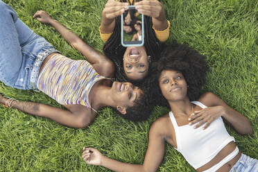 Fröhliche Frau nimmt Selfie durch Handy mit weiblichen Freunden auf Gras - JCCMF03925