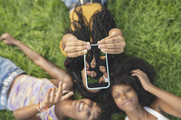 Junge Frau, die ein Selfie mit ihren Freundinnen macht, während sie im Gras liegt - JCCMF03926