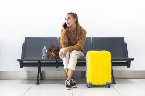 Woman with wheeled luggage talking on mobile phone in airport - JAQF00782