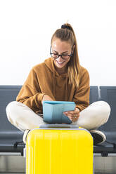 Woman using digital tablet while sitting on bench in airport - JAQF00786