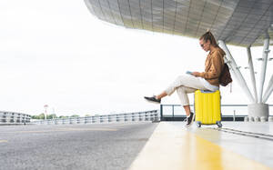 Junge Frau, die ein digitales Tablet benutzt, während sie auf einem Rollkoffer am Flughafen sitzt - JAQF00795