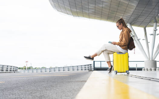 Junge Frau, die ein digitales Tablet benutzt, während sie auf einem Rollkoffer am Flughafen sitzt - JAQF00795