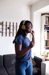 Smiling mid adult woman listening music through wireless headphones while dancing in living room - ASGF01538