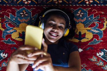 Glückliche Frau spricht Selfie durch Handy, während auf Teppich im Wohnzimmer liegen - ASGF01541