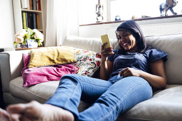 Smiling woman surfing net through smart phone while sitting on sofa at home - ASGF01548