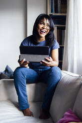 Fröhliche Frau mit digitalem Tablet, die zu Hause auf dem Sofa sitzt - ASGF01551