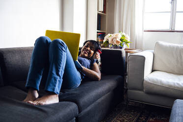 Lächelnde Frau im mittleren Erwachsenenalter, die einen Laptop benutzt, während sie auf dem Sofa im Wohnzimmer liegt - ASGF01566
