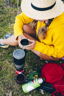Mittlere erwachsene Frau mit Hut, die eine Tasse hält, während sie am Campingkocher sitzt - OMIF00078
