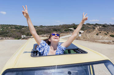 Glückliche Frau mit Friedenszeichen, die sich im Sommer durch das Schiebedach eines Lieferwagens lehnt - PGF00822