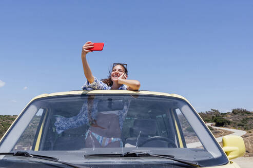 Glückliche Frau, die ein Selfie mit ihrem Smartphone macht, während sie sich durch das Schiebedach eines Vans lehnt - PGF00826