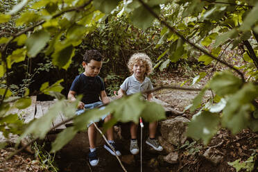 Boy talking with male friend while sitting on footbridge in forest - MRRF01527