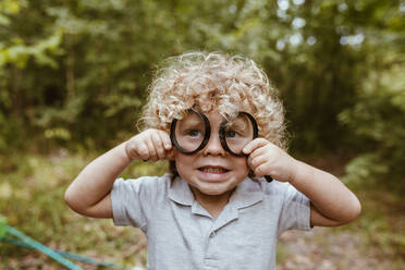 Junge mit lockigem Haar, der im Wald durch ein Vergrößerungsglas schaut - MRRF01538