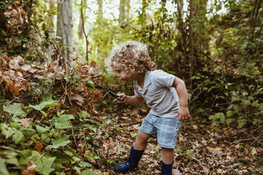 Neugieriger Junge mit lockigem Haar betrachtet Pflanzen durch ein Vergrößerungsglas im Wald - MRRF01548