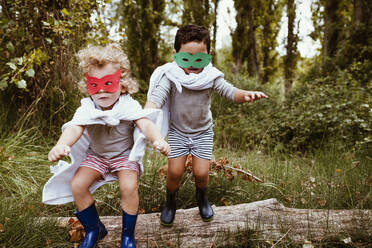 Verspielte Jungen mit Masken und Umhängen, die zusammen im Wald springen - MRRF01567