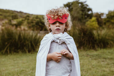 Cute blond boy with curly hair wearing mask and cape standing on meadow - MRRF01574