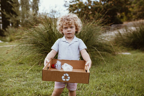 Blonder Junge mit lockigem Haar trägt Karton mit Plastik auf einer Wiese - MRRF01578