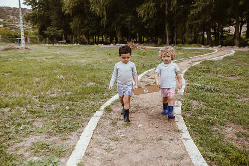 Jungen tragen wiederverwertbare Pappkartons, während sie gemeinsam auf einem Weg gehen - MRRF01579