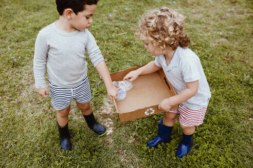Junge schaut auf männlichen Freund, der Plastik in einem wiederverwertbaren Karton auf der Wiese aufbewahrt - MRRF01581