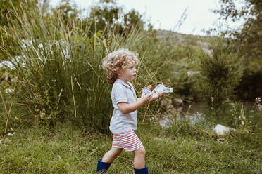 Blond boy with curly hair carrying plastics while walking on meadow - MRRF01582