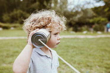 Cute blond boy with curly hair listening through tin can phone on meadow - MRRF01585