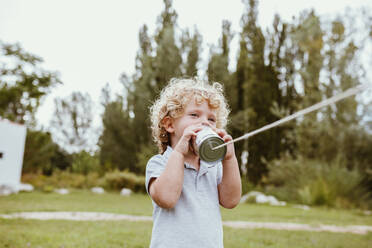Boy with blond curly hair talking through tin can phone on meadow - MRRF01587