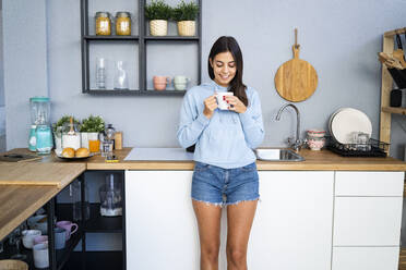 Smiling woman holding coffee mug while standing by kitchen counter - GIOF13583